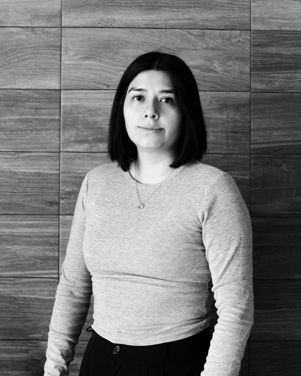 Tania Muñoz de pie frente a un muro de madera, con suéter claro y el cabello a la altura de la mandíbula. Fotografía sin color.