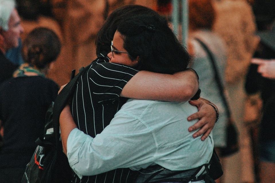 Dos personas se abrazan de pie, entre gente, con la luz cálida del final de la tarde.